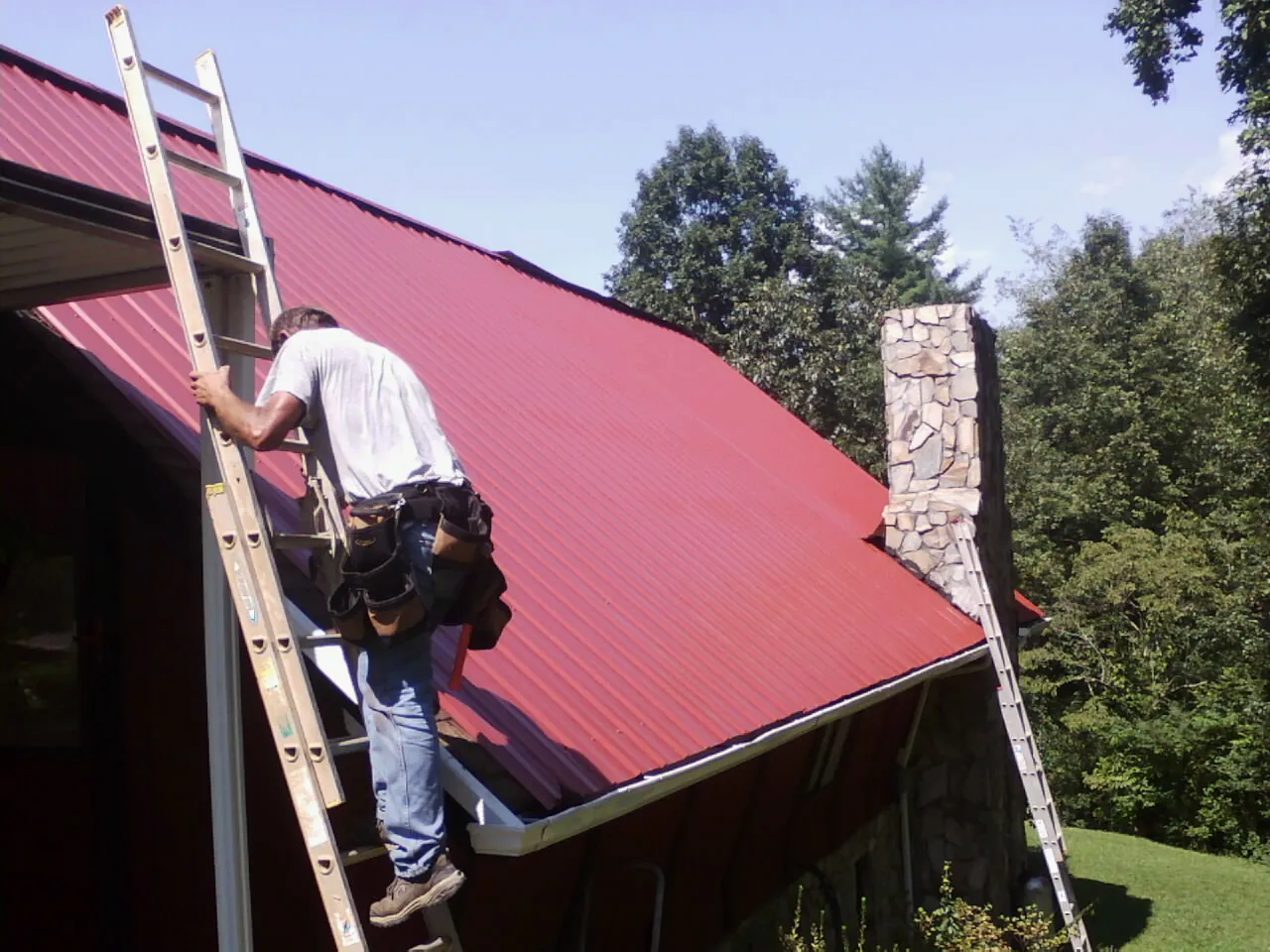 Roofer on a ladder beside a red metal roof and stone chimney — Soffit & Fascia Repair by Roofing Brothers in Signal Mountain